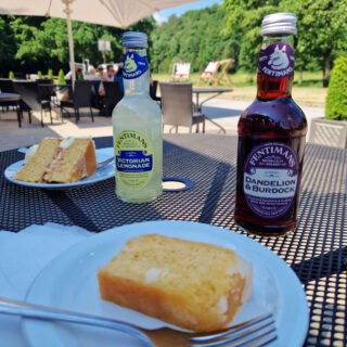 In the foreground is a slice of lemon cake with a fork on a white plate on a mesh table. Behind is a bottle of brown drink, and next to it a bottle of yellow drink and a triangular slice of cake on a plate. The table is outside a cafe in the countryside
