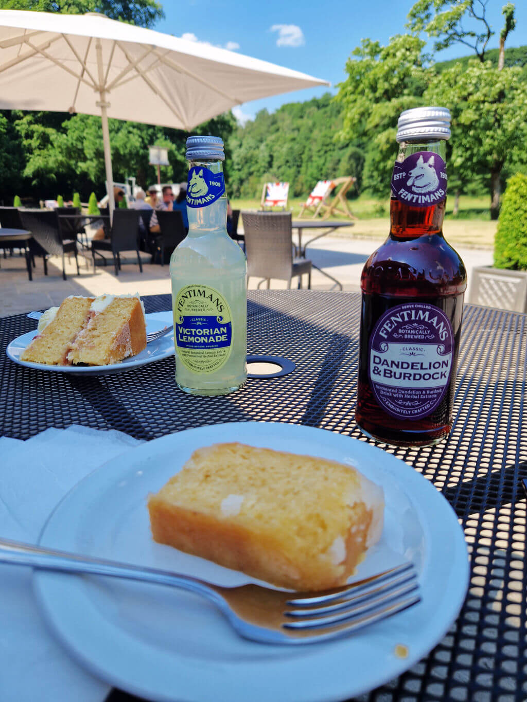 In the foreground is a slice of lemon cake with a fork on a white plate on a mesh table. Behind is a bottle of brown drink, and next to it a bottle of yellow drink and a triangular slice of cake on a plate. The table is outside a cafe in the countryside