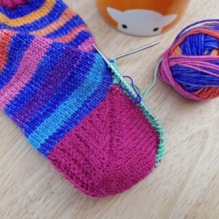 A completed Fish Lips Kiss sock heel. The leg of the sock is blue and rainbow stripes and the heel is pink. The sock is lying on a wooden table next to an orange mug of tea