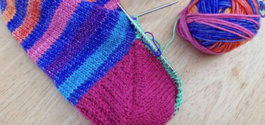 A completed Fish Lips Kiss sock heel. The leg of the sock is blue and rainbow stripes and the heel is pink. The sock is lying on a wooden table next to an orange mug of tea