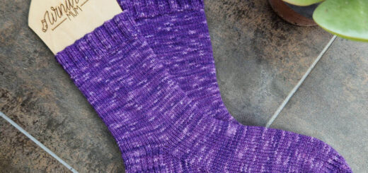 A pair of purple variegate socks on wooden sock blockers inscribed with the Winwick Mum logo are lying on a brown tiled floor next to a succulent plant