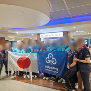 A group of young women wearing tuquoise hoodies and navy blue t-shirts standing in a group holding a Japanese flag and a Girl Guiding flag. Their faces have been blurred for privacy.