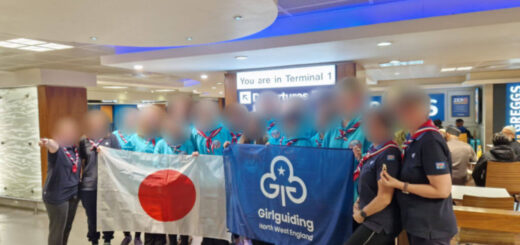 A group of young women wearing tuquoise hoodies and navy blue t-shirts standing in a group holding a Japanese flag and a Girl Guiding flag. Their faces have been blurred for privacy.