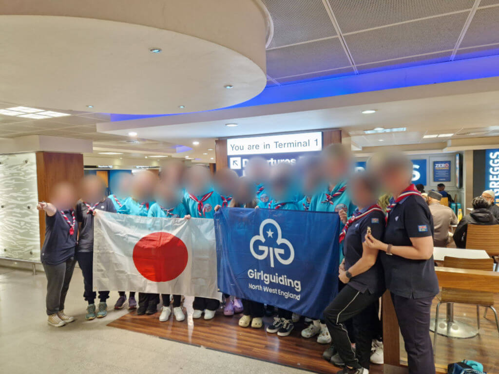A group of young women wearing tuquoise hoodies and navy blue t-shirts standing in a group holding a Japanese flag and a Girl Guiding flag. Their faces have been blurred for privacy.