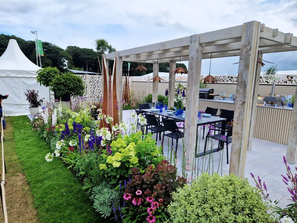 A weathered wood trellis on a patio providing cover and shade for a table laid for dinner. The planted border runs alongside the patio. RHS Tatton Show 2023