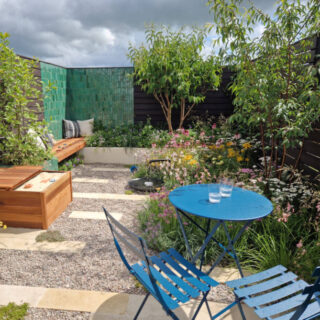 A family garden space bordered by a green tiled wall and dark brown fence. The planting is around the edges of the garden, a children's sand box is against one wall and a blue metal table and chairs closest to the camera
