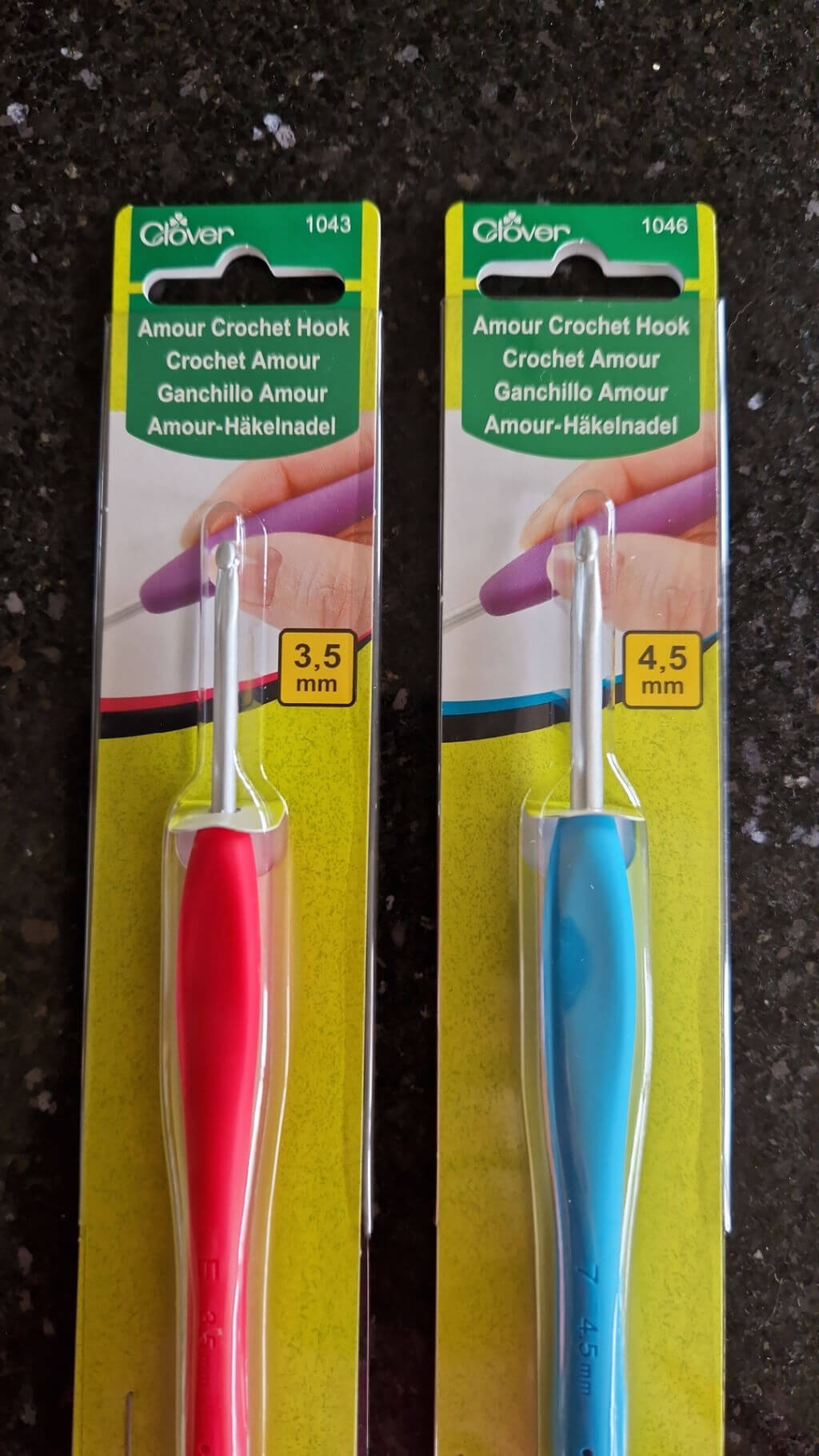 Two crochet hooks in different sizes, still in their packaging, lie on a granite worktop. The left hand hook has a red handle and the right hand hook has a blue handle.