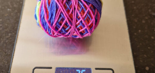 A ball of multi-coloured yarn on a flat silver-coloured weighing scale. The ball of yarn weighs 25g.