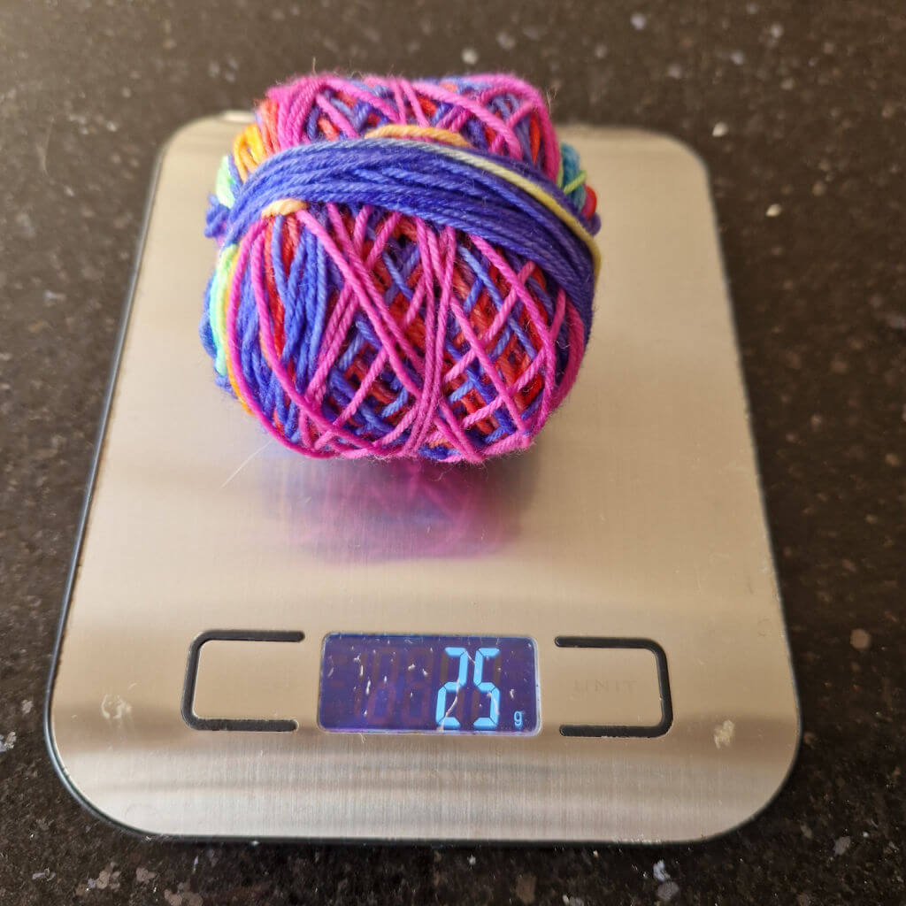A ball of multi-coloured yarn on a flat silver-coloured weighing scale. The ball of yarn weighs 25g.