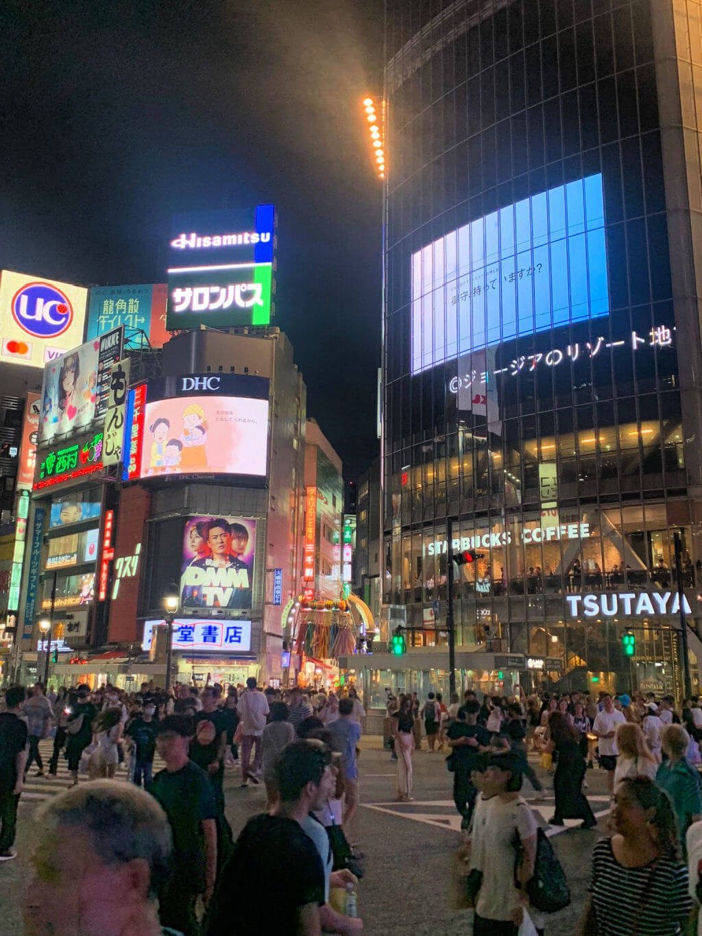 A crowd of people on a road crossing in a busy city. The crossing is surrounded by tall buildings with advertising screens on them