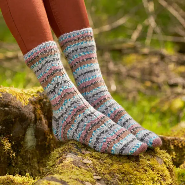 A pair of pink, grey, blue, black and white socks modelled on feet. The wearer is standing on a mossy log.