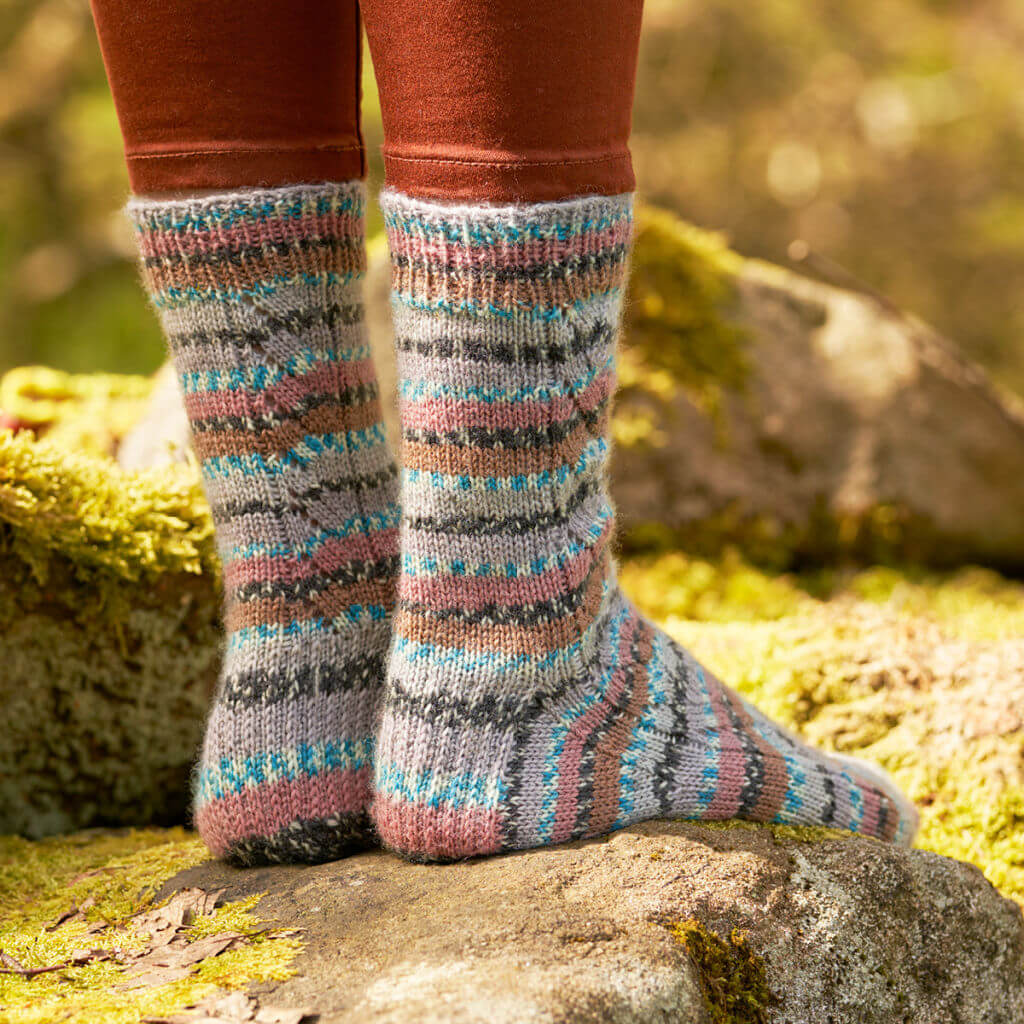 A pair of pink, grey, blue, black and white socks modelled on feet. The pattern panel down the ack of the sock is visible. The wearer is standing on a mossy log.