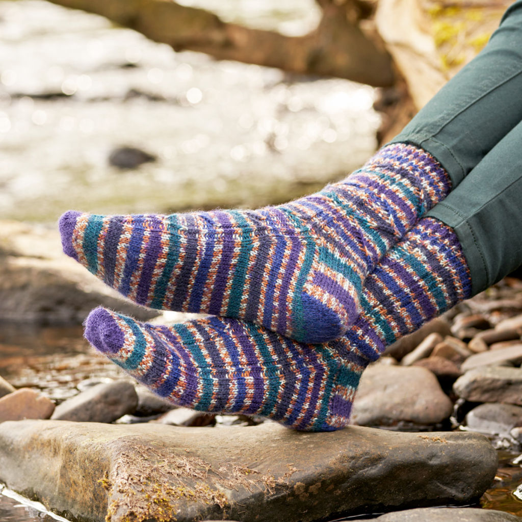 A pair of navy, teal, purple, gold and white socks modelled on feet. The wearer is standing on a mossy log.