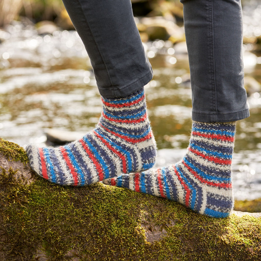 A pair of orange, navy, blue, black and white socks modelled on feet. The wearer is standing on a mossy log.