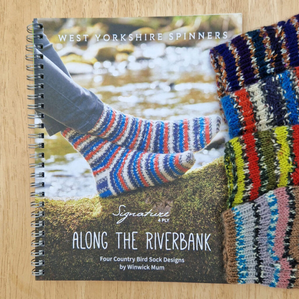 A pattern book with a pair of red, orange, white and black striped socks on the front. To the right, four pairs of socks are lined up against the edge of the book, only their cuffs showing