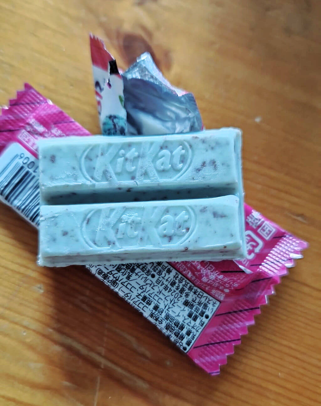 A mini KitKat biscuit lying on a pink wrapper on a wooden table. The KitKat is the same colour as mint choc chip ice cream