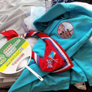 An open suitcase with a turquoise blue hoody, a red, blue and white Guide necker (neckerchief) and a green and yellow fan lying on it