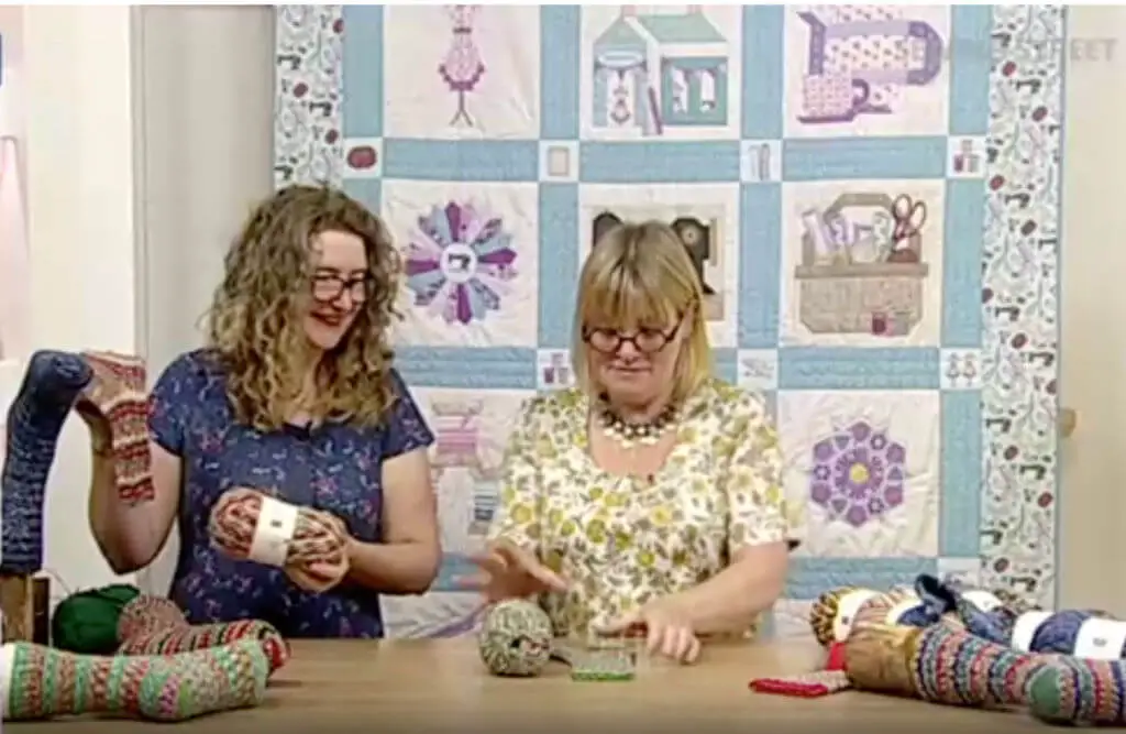 Two women standing at a table discussing knitted socks and yarn