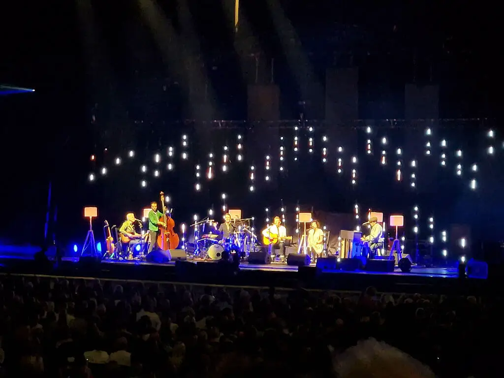 A band on a stage, all seated and with standard lamps surrounding them to light up the set