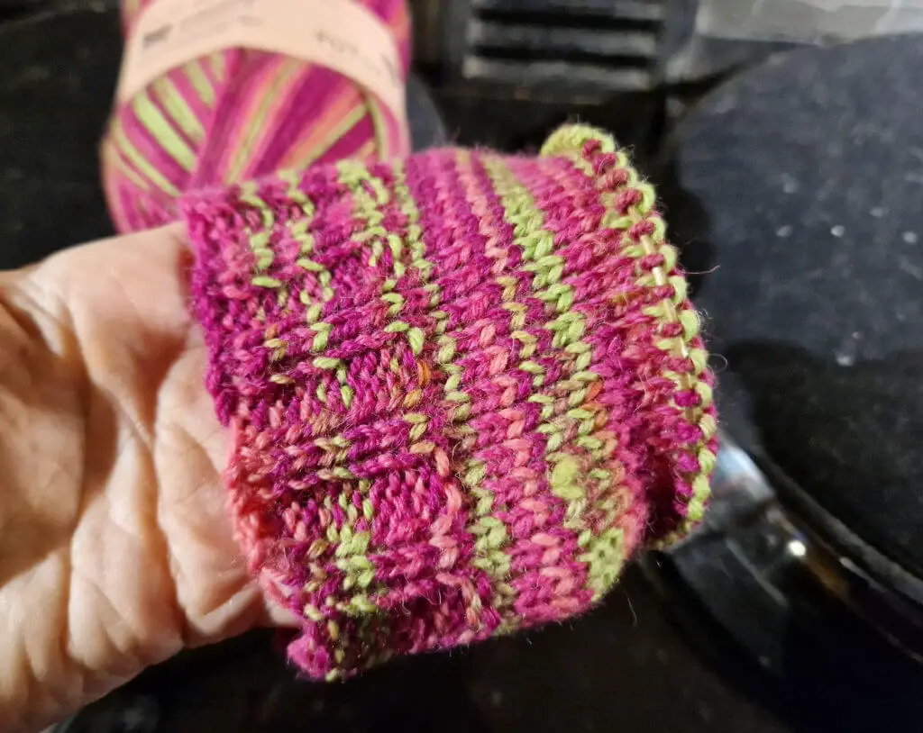 A partly knitted sock held so that you can see the pink and neon green stripes