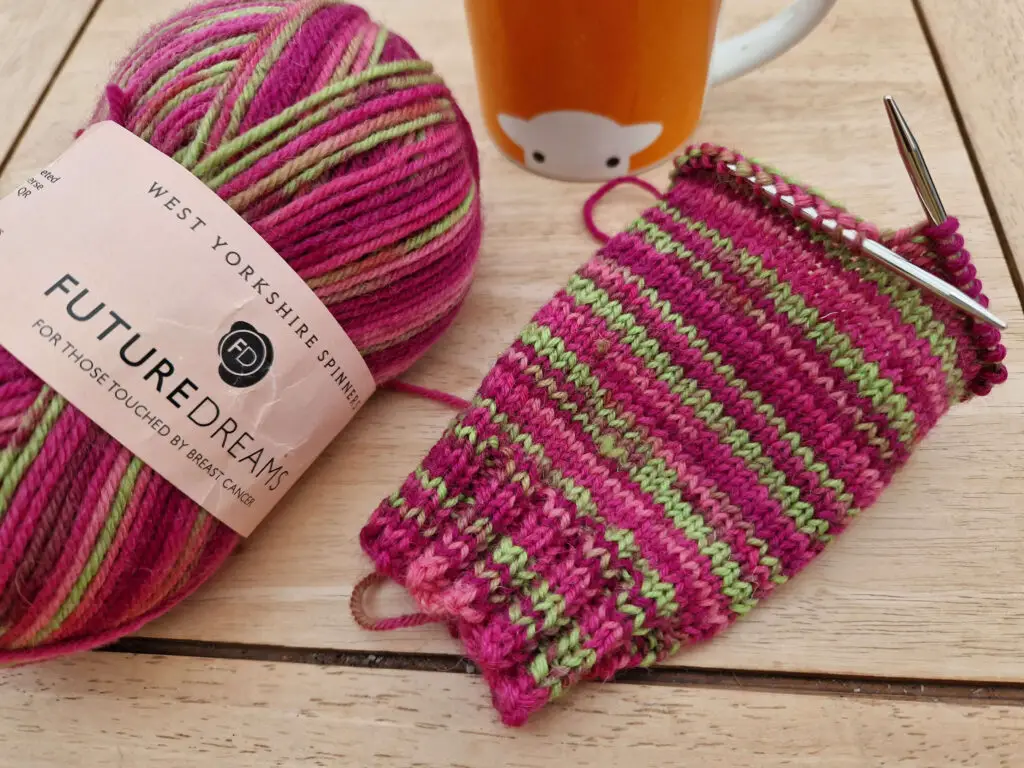 A partly knitted sock in stripes of pink and neon green is lying on a wooden table next to the ball of yarn and an orange mug
