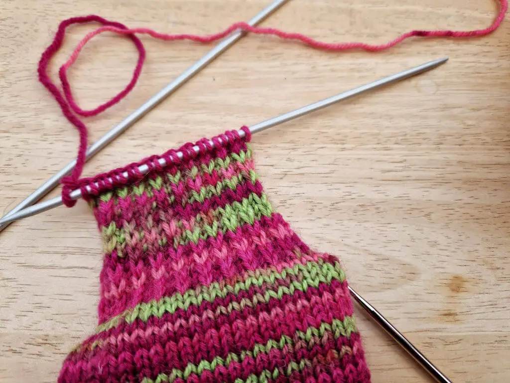 The heel flap of a partly knitted sock on a double pointed knitting needle, lying on a wooden table