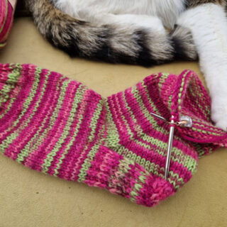 A partly-knitted pink and green sock on a mustard sofa. A tabby and white cat has one paw on the sock