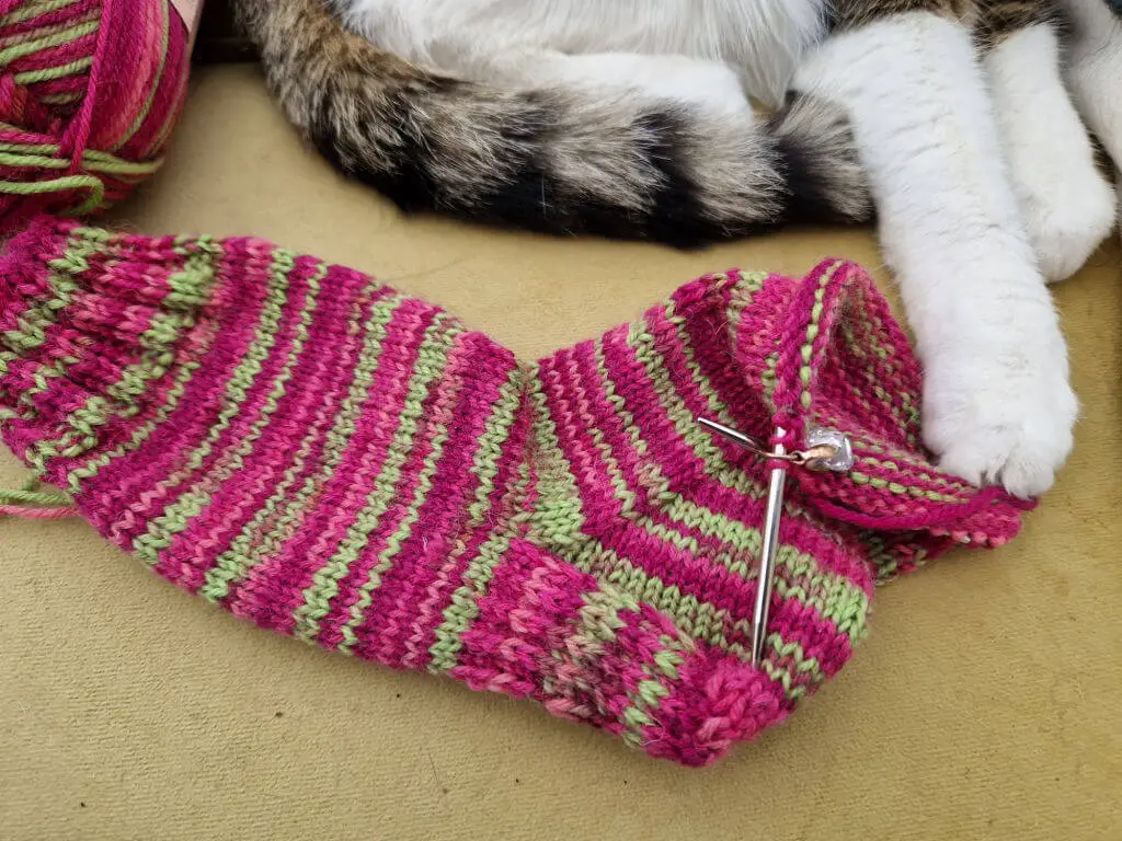 A partly-knitted pink and green sock on a mustard sofa.  A tabby and white cat has one paw on the sock