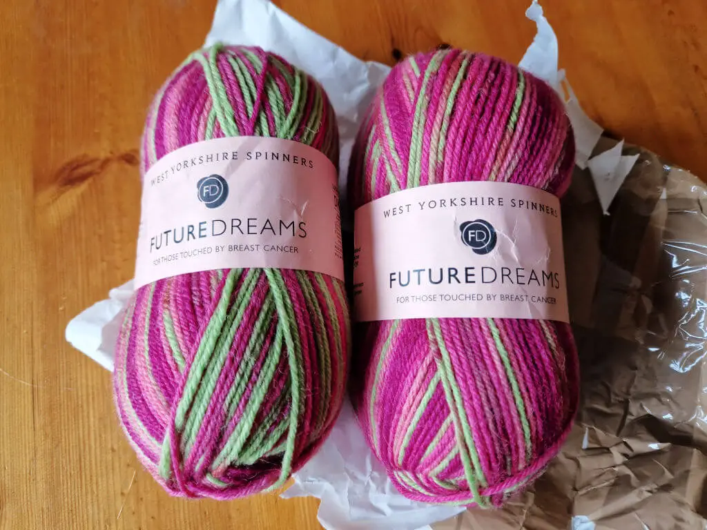 Two balls of pink and neon green yarn lying on the postal packing they came in, on a wooden table