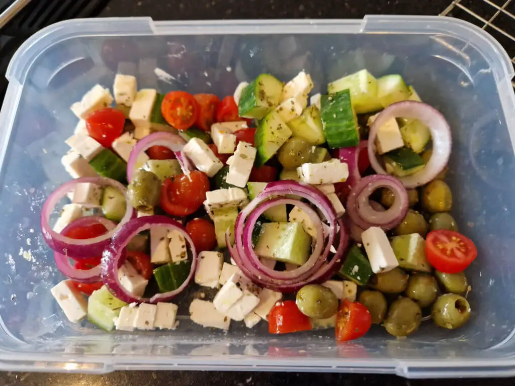 A clear plastic storage container filled with Greek salad containing tomatoes, cucumber, olives, feta cheese and onions