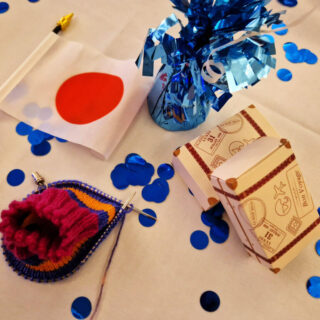A partly-knitted striped sock, a Japanese flag, two cardboard favours boxes in the shape of suitcases and a blue foil balloon weight are on a white table cloth with blue shiny confetti circles scattered around them