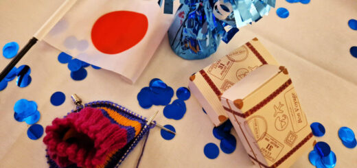 A partly-knitted striped sock, a Japanese flag, two cardboard favours boxes in the shape of suitcases and a blue foil balloon weight are on a white table cloth with blue shiny confetti circles scattered around them