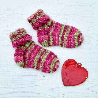 A pair of baby socks in pink and green stripes with a decorative cuff lie on a white background next to a red glass heart