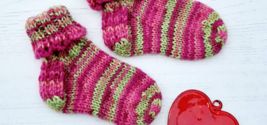 A pair of baby socks in pink and green stripes with a decorative cuff lie on a white background next to a red glass heart