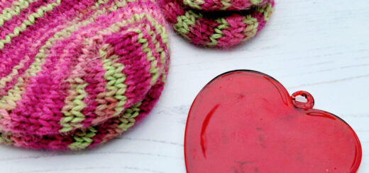 Two pairs of pink and green striped socks - one adult, one baby, on a white wooden table with a red glass heart