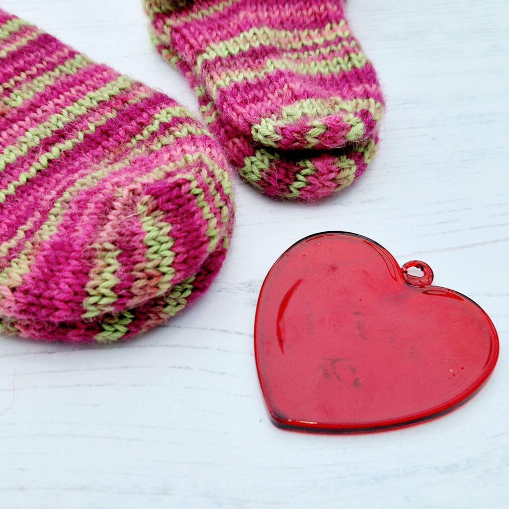 Two pairs of pink and green striped socks - one adult, one baby, on a white wooden table with a red glass heart