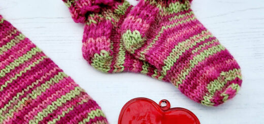 A pair of baby socks in pink and green stripes with a decorative cuff lie on a white background next to an adult sock in the same yarn and a red glass heart