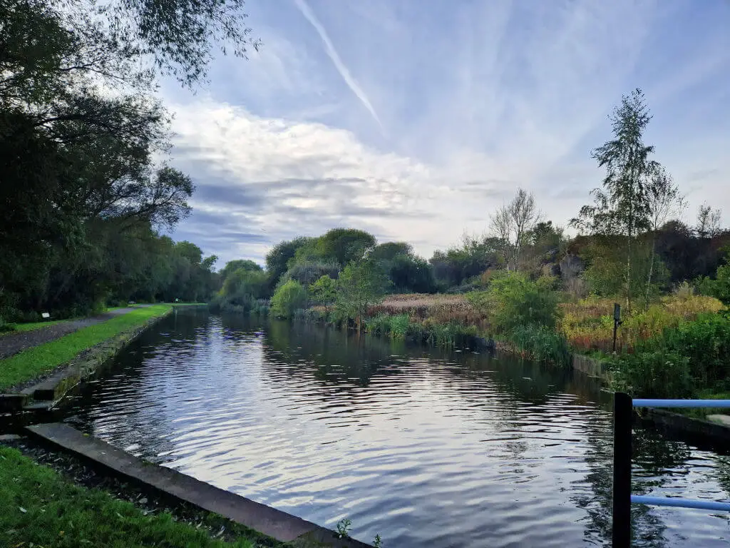 A view across a canal surrounded by trees and bushes. The sky above is blue streaked with clouds