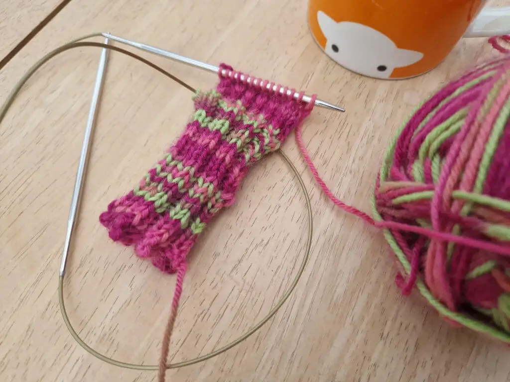 A partly-knitted sock on a long circular knitting needle lying on a wooden table next to a ball of pink and green yarn and an orange mug