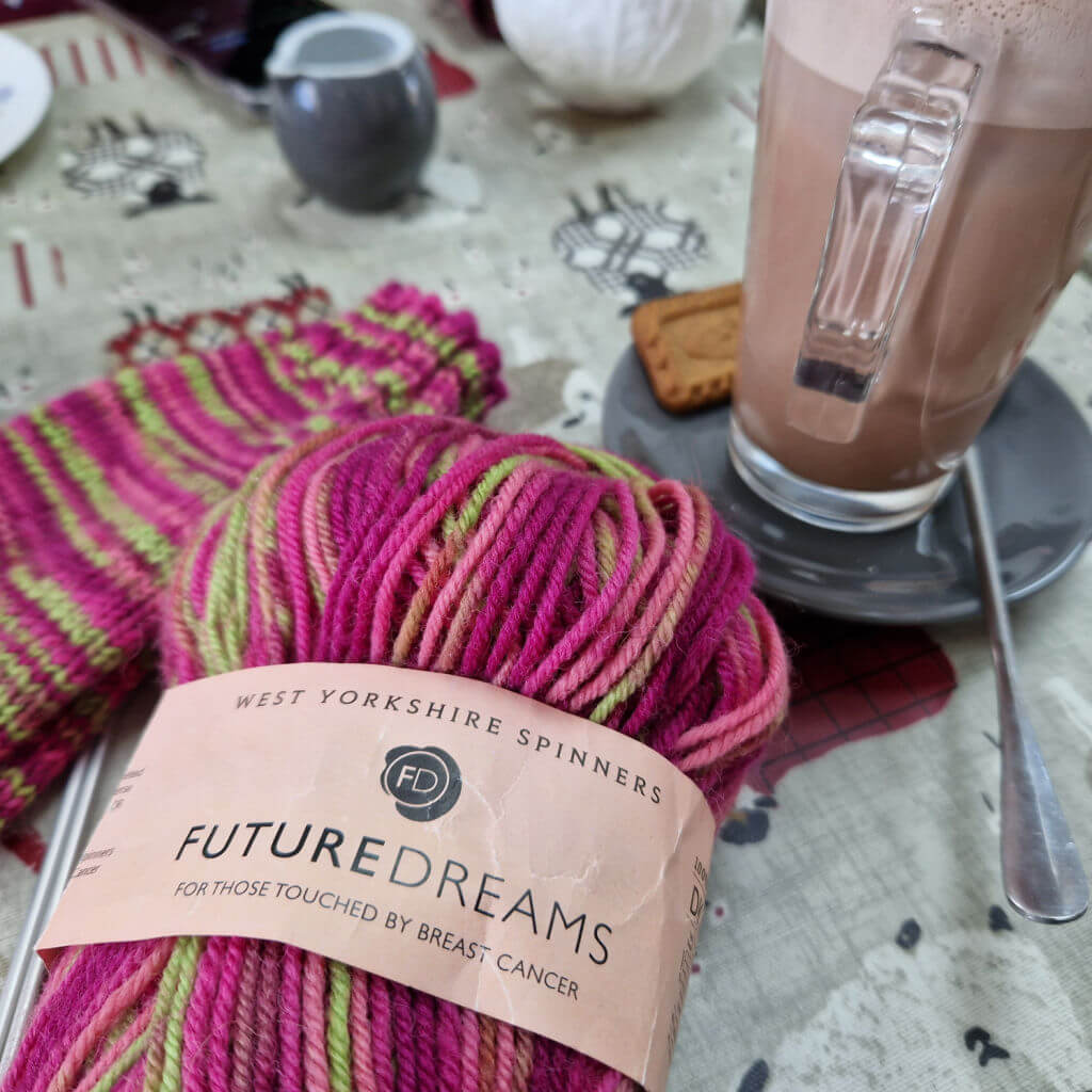 A cafe table with a pink and green striped sock and ball of yarn on it, and a glass of hot chocolate in a saucer.