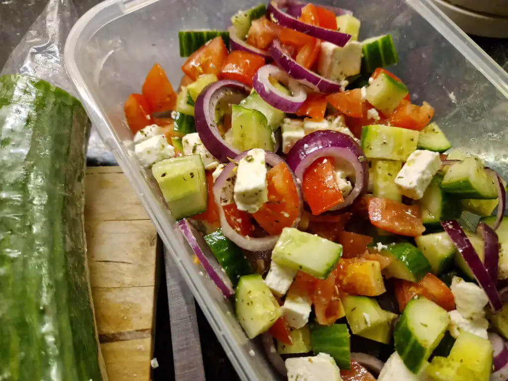 A storage box of Greek salad next to a cucumber on a working top