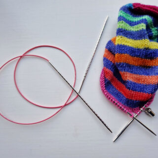 A partly-knitted sock in stripes of blue and rainbow on a white background next to a curled long circular needle with a pink cable