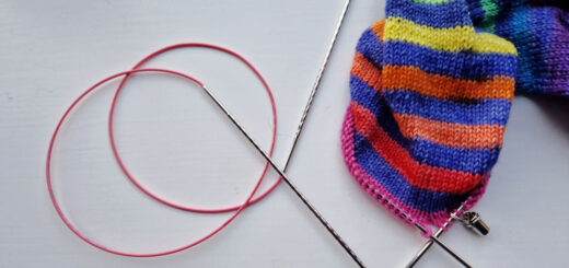 A partly-knitted sock in stripes of blue and rainbow on a white background next to a curled long circular needle with a pink cable