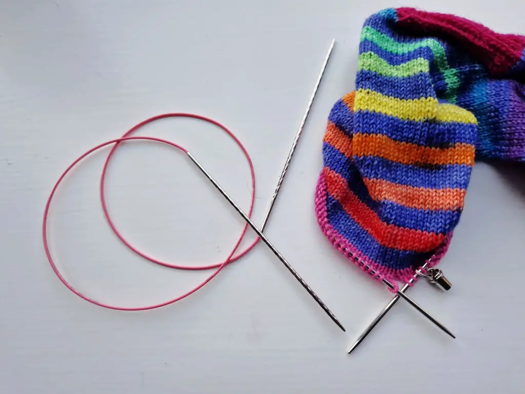 A partly-knitted sock in stripes of blue and rainbow on a white background next to a curled long circular needle with a pink cable