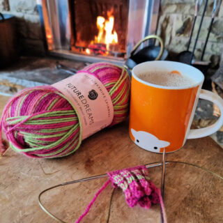 A partly-knitted sock on a long circular knitting needle lying on a wooden table next to a ball of pink and green yarn and an orange mug in front of an open fire