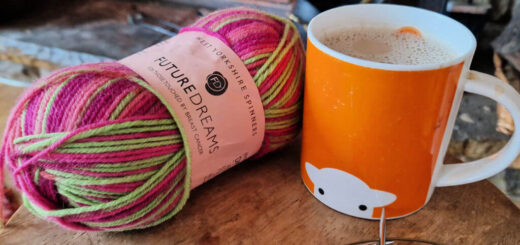 A partly-knitted sock on a long circular knitting needle lying on a wooden table next to a ball of pink and green yarn and an orange mug in front of an open fire