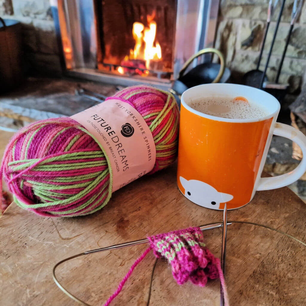A partly-knitted sock on a long circular knitting needle lying on a wooden table next to a ball of pink and green yarn and an orange mug in front of an open fire