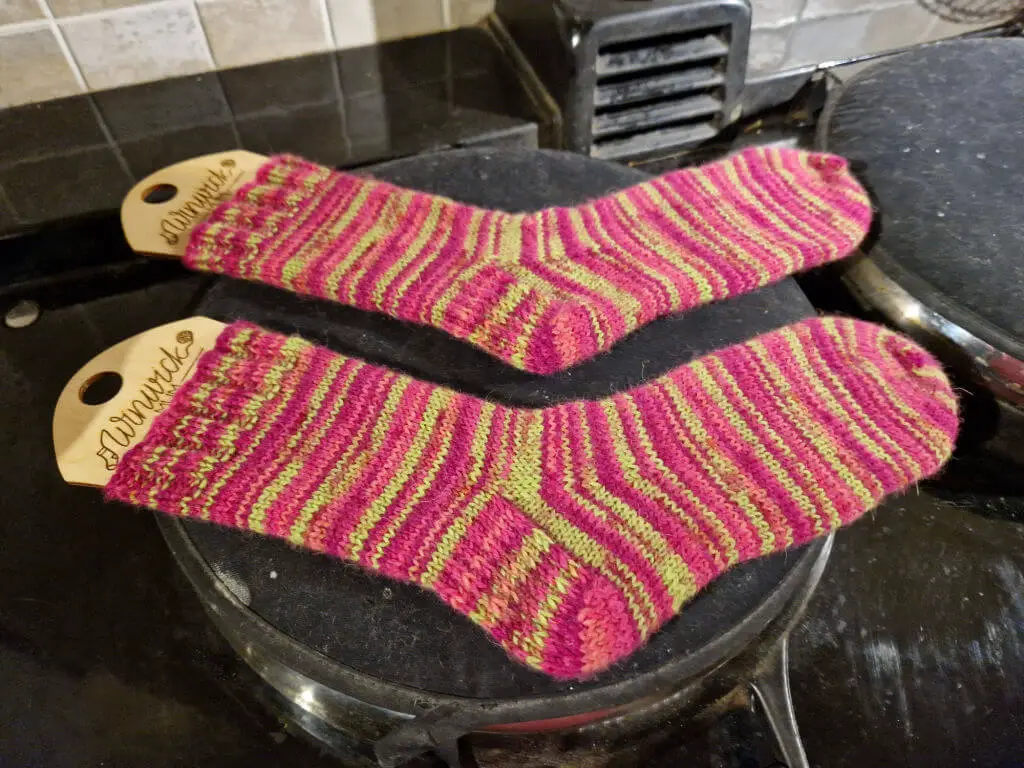 A pair of pink and green striped socks on wooden sock blockers drying on an Aga lid
