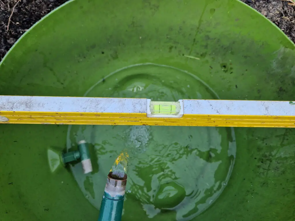 A spirit level balanced across the base of an old water barrel cut to make a small pond. The pond is being filled up with water