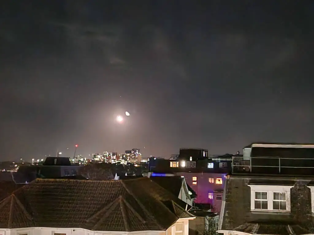 A view across roof tops taken at night. The moon is bright and there are lights on the buildings in the distance.
